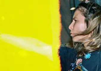 Person with light hair in a bun, wearing a graphic shirt, in profile beside a vivid yellow barrier/wall.