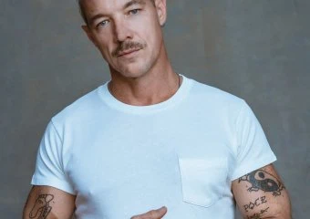 Man with short blond hair and tattoos on both arms, wearing a light blue T-shirt, arms crossed against a gray backdrop.
