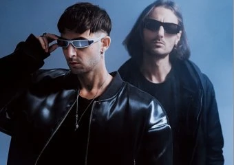 Two men wearing sunglasses and black leather jackets pose against a blue background.