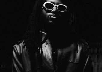 Person with dreadlocks wearing sunglasses and a leather jacket in low light.