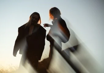 Two people in dark coats stand outdoors at sunset, with strong backlight and motion blur.