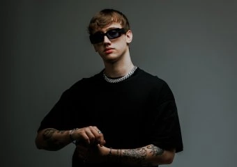 Young man in a black t-shirt with dark sunglasses and a chunky silver chain, tattoos visible on both arms, against a dark gray background.