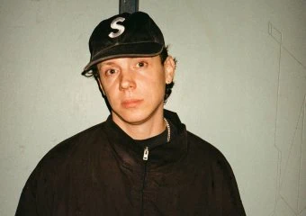 Young man wearing a black jacket and cap, standing against a plain wall.