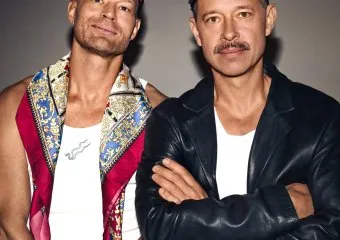 Two men posing, one with a colorful scarf and sunglasses, the other with a black cap and leather jacket.