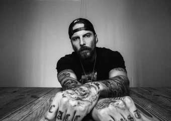 Black-and-white portrait of a tattooed man leaning on a wooden table, wearing a backward cap and chain necklace.