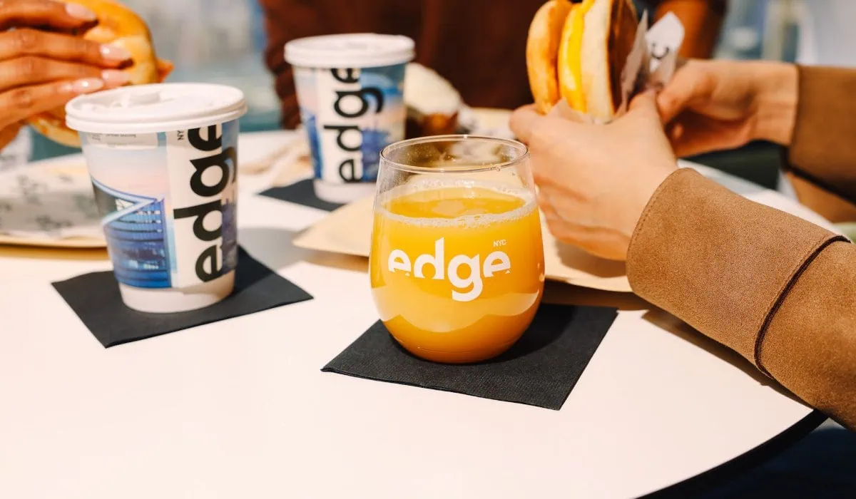 Orange juice in a glass with the 'edge NYC' branding on a white table, surrounded by sandwiches and branded cups.