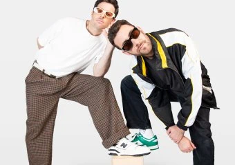 Two men with sunglasses posing on wooden blocks against a white background.