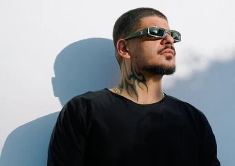 Man wearing a black shirt and sunglasses stands against a white wall, showing a bold neck tattoo and a mesh-covered forearm with bracelets.