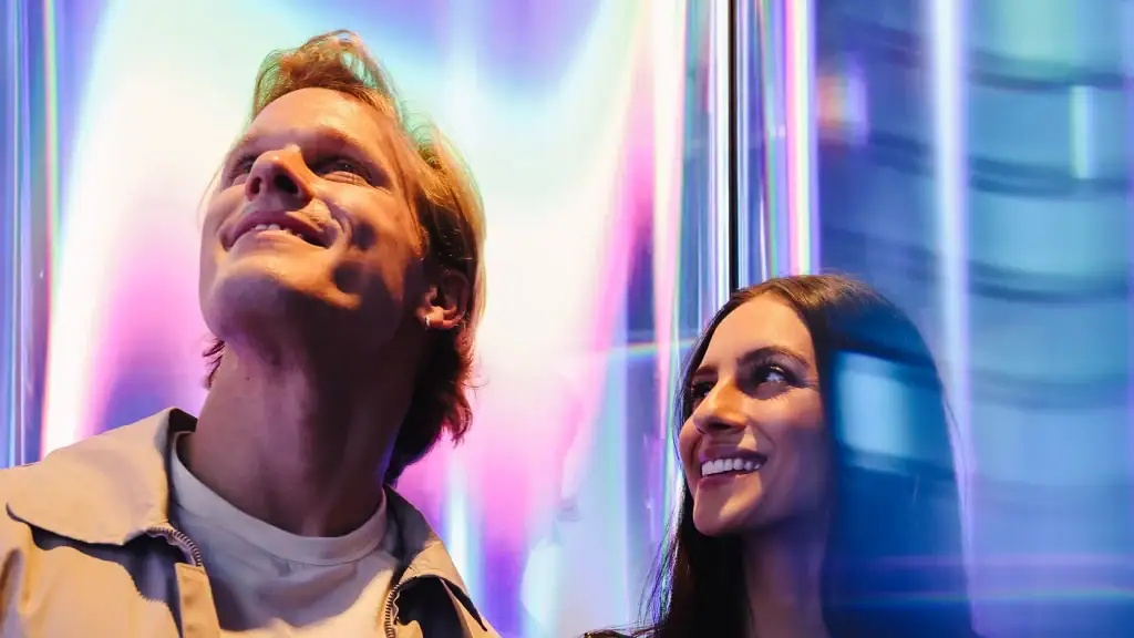 man-woman-at-skyrise-immersive-experience-at-edge - Edge NYC - Edge NYC Two friends stand close together, smiling at colorful neon lights behind them in a reflective, purple-blue glow.
