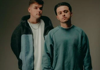 Two young men stand side by side in casual outfits against a dark gradient backdrop in a studio setting.