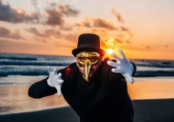 Person in a gold masquerade mask and black top hat on a sunset beach, reaching toward the camera with white gloves.