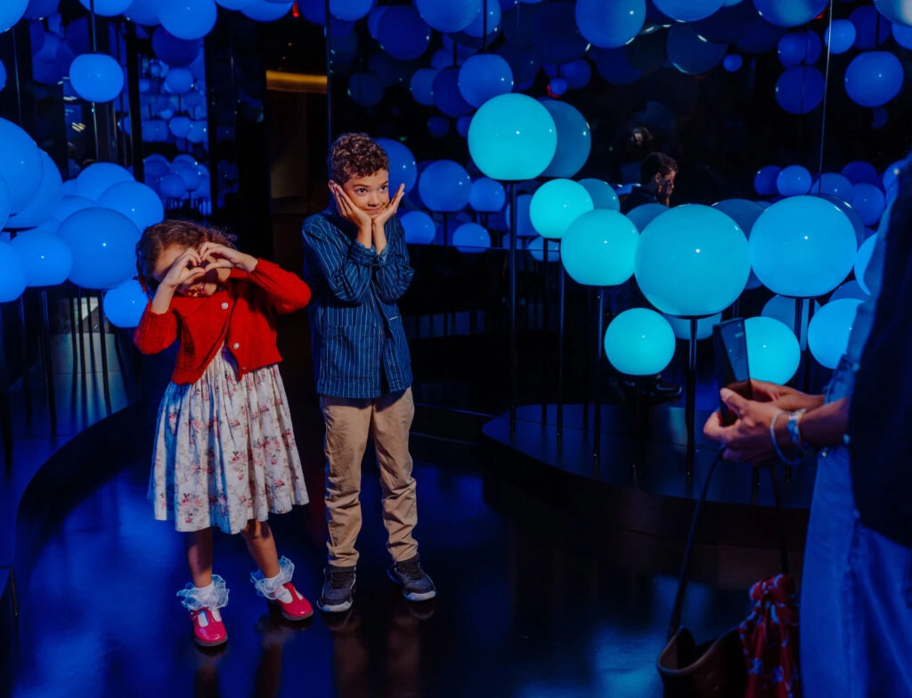 Edge NYC Indoor Art Installation with Children - Edge NYC - Edge NYC Two children pose with hands on their cheeks among blue illuminated balloons in a mirrored room.