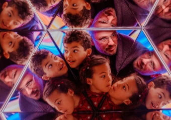 Children and adults lean in around a mirrored, geometric kaleidoscope, looking up with smiles amid colorful lights.