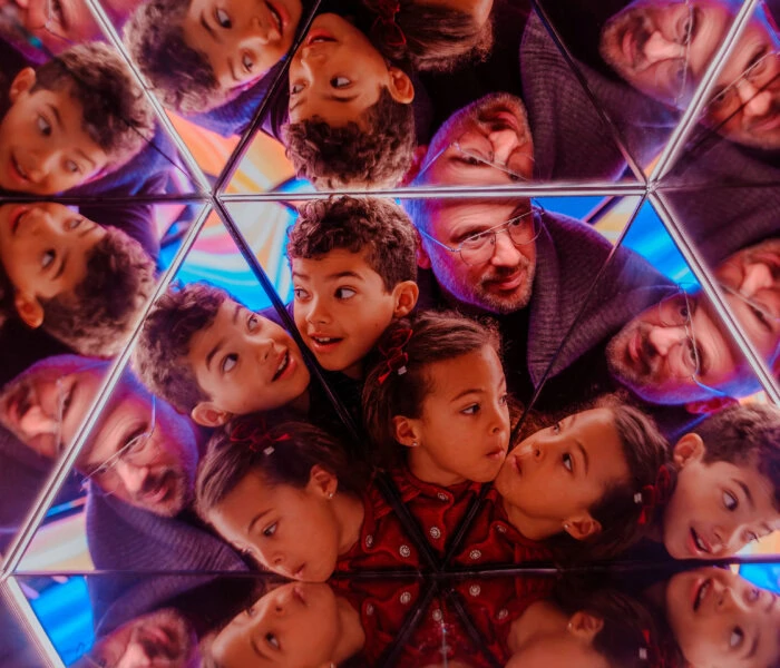 Children and adults lean in around a mirrored, geometric kaleidoscope, looking up with smiles amid colorful lights.
