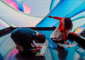 Two children reading colorful brochures on a shiny, abstract blue-pink floor in a bright exhibit far wall concept art surface.