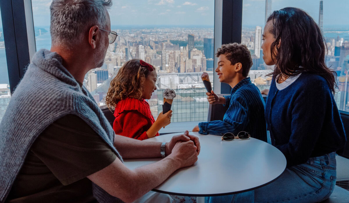 A family of four sits around a round table indoors, each holding ice cream cones, with a city skyline visible through large windows behind them.