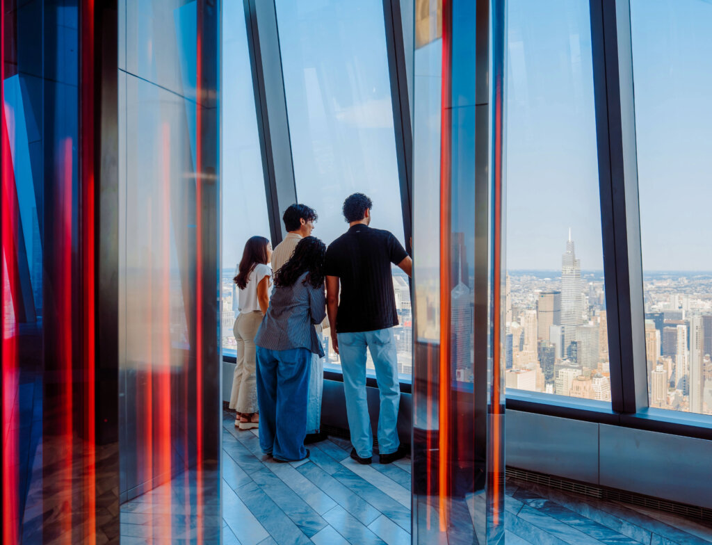 2026-Edge-MediabyAV-InfiniteCity1 1 - Edge NYC - Edge NYC Four people stand together in a glass-walled viewing area, looking out over a city skyline from a high-rise.