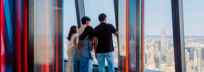 Four people stand together in a glass-walled viewing area, looking out over a city skyline from a high-rise.