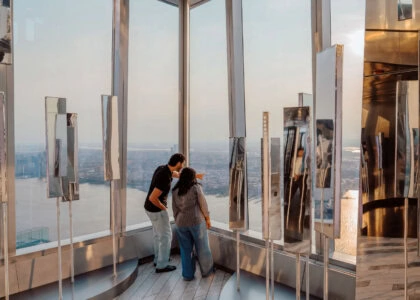 Two people stand at a glass-walled observation deck, gazing out at a distant city skyline.