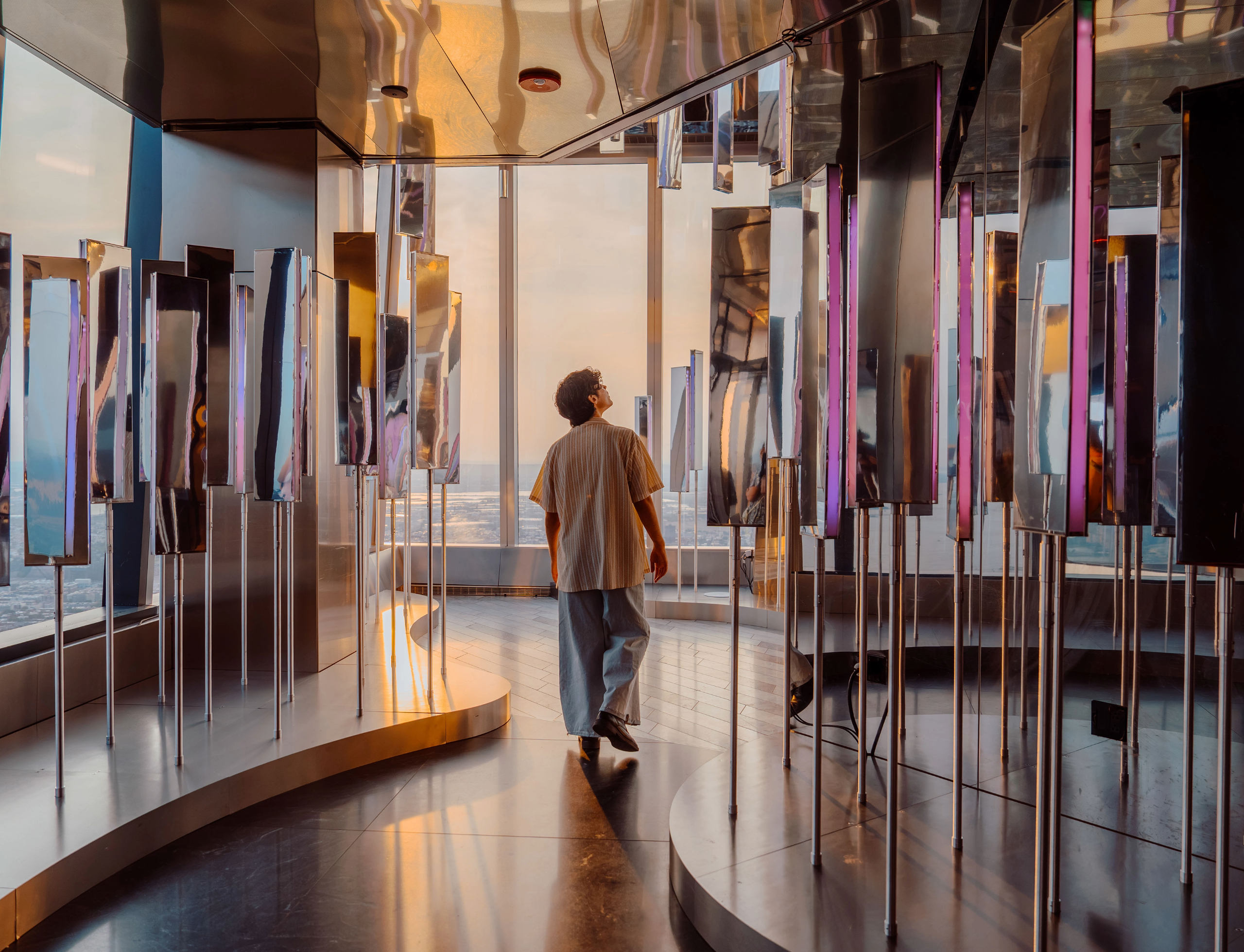 Person in a striped shirt walks through a foyer of tall, reflective mirrored panels lit by warm sunset light.