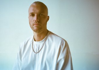 Close-up portrait of a man with short hair and a chain necklace, neutral background.