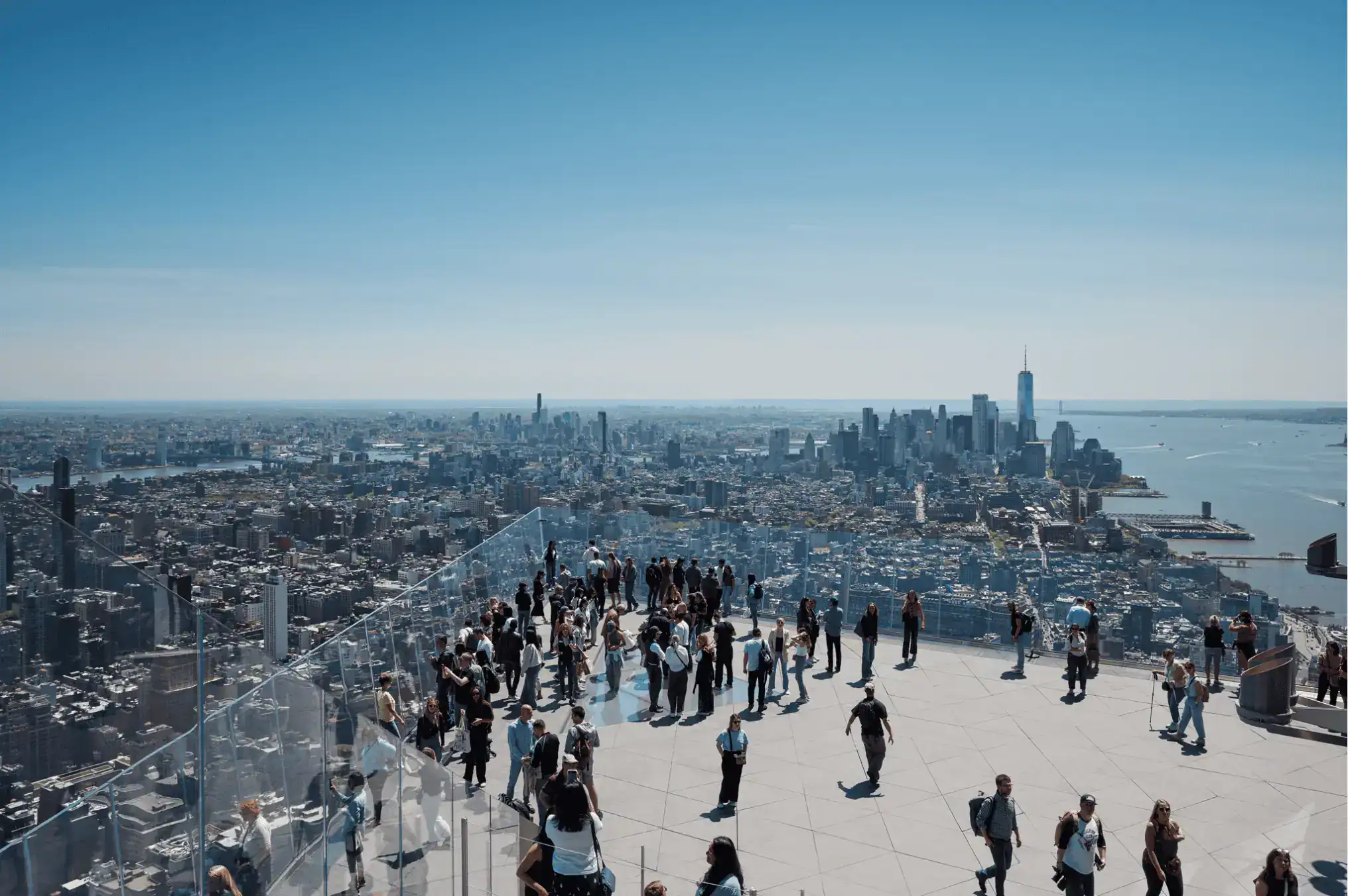 Crowded outdoor observation deck with glass railing overlooking the New York City skyline and river in the distance.