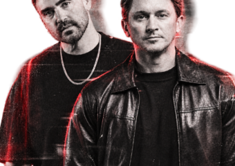 Two men pose for a promo photo: one in a black leather jacket in front, the other in a black T-shirt with a chain behind him, both under red lighting.
