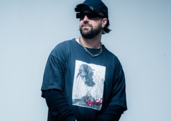 Bearded man in a blue graphic T-shirt, dark cap and sunglasses, standing against a blue gradient wall.