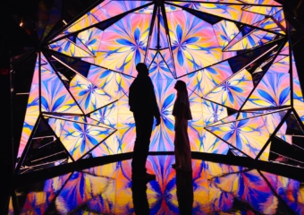 Silhouetted man and woman stand before a vibrant geometric light installation with pink, yellow, and blue panels reflected on a glossy floor.