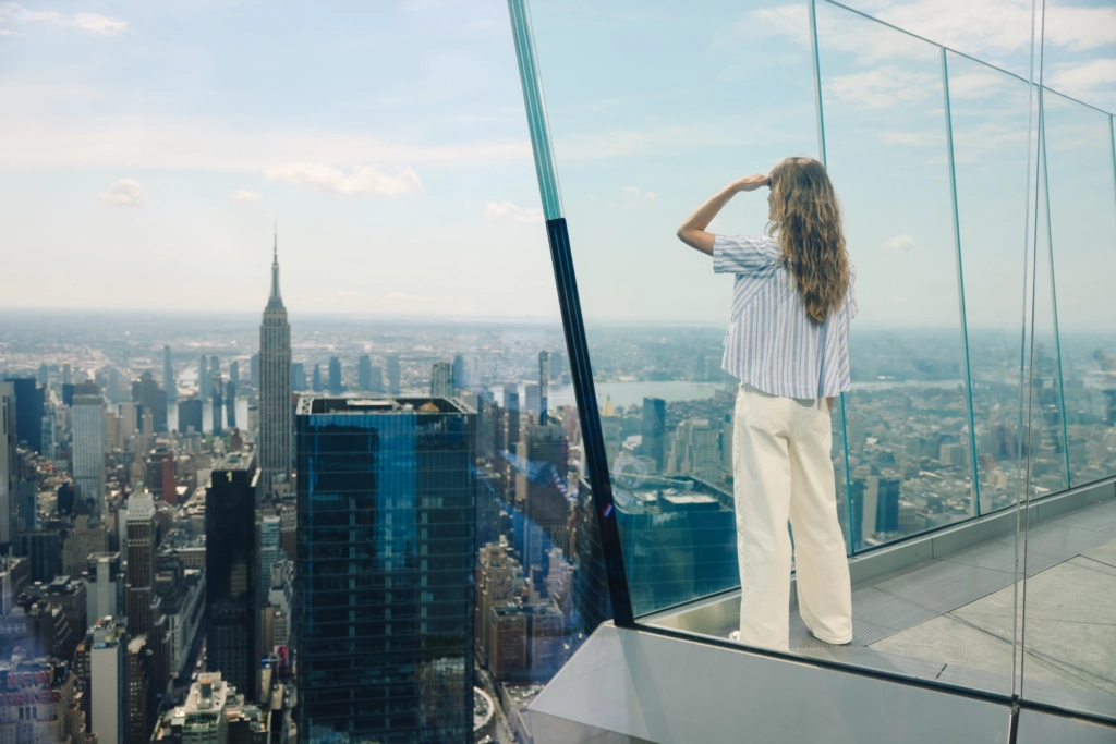 Woman enjoying panoramic NYC skyline view at Edge - Edge NYC - Edge NYC Person with long hair stands on a glass observation deck, looking over the New York City skyline.