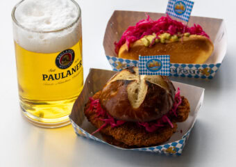 Beer in a Paulaner glass alongside two pretzel buns with pink cabbage on blue-white checkered boxes, on a white table.