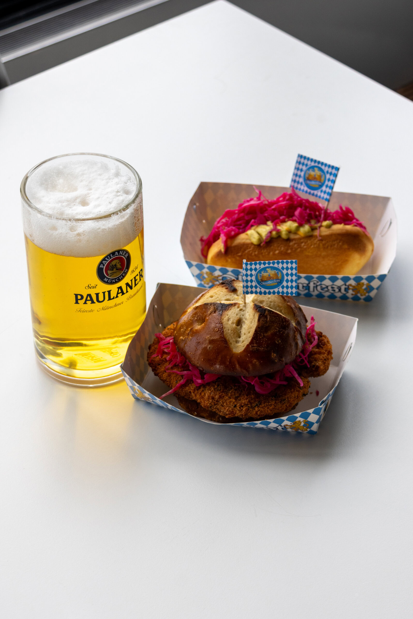 Beer in a Paulaner glass alongside two pretzel buns with pink cabbage on blue-white checkered boxes, on a white table.