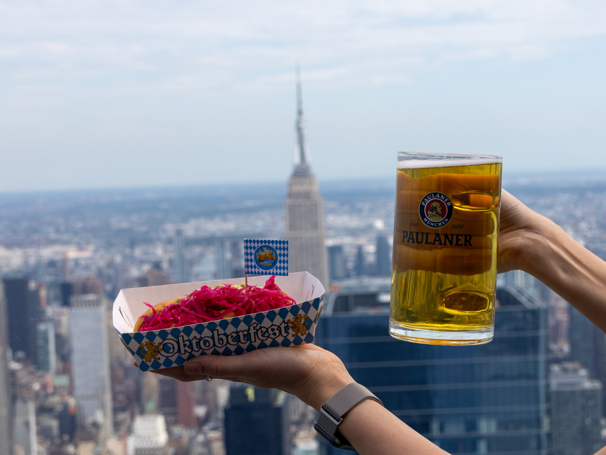 Edge NYC Sky Deck with Oktoberfest Beer - Edge NYC - Edge NYC A glass of Paulaner beer being held up on a rooftop with a blurry city skyline in the background and an Oktoberfest-themed box in the foreground.