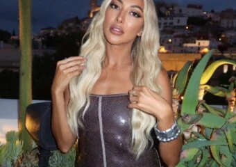 Woman with long blonde hair posing in a sparkly dress on a terrace, night city lights in the background among cacti and plants.