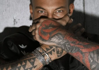 Close-up of a tattooed man covering his mouth with crossed arms, wearing a black jacket against a concrete wall.
