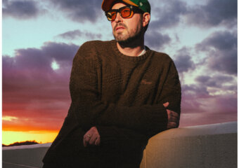Man leaning on a concrete wall at sunset, wearing a brown knit sweater, green cap, and orange sunglasses against a colorful sky.