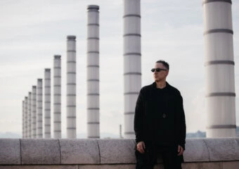 Man in a black coat and sunglasses stands beside a row of tall white industrial smokestacks on a rooftop.