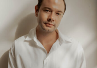 Portrait of a man in a white button-down shirt leaning slightly against a neutral wall with a soft, warm light and a gentle smile.